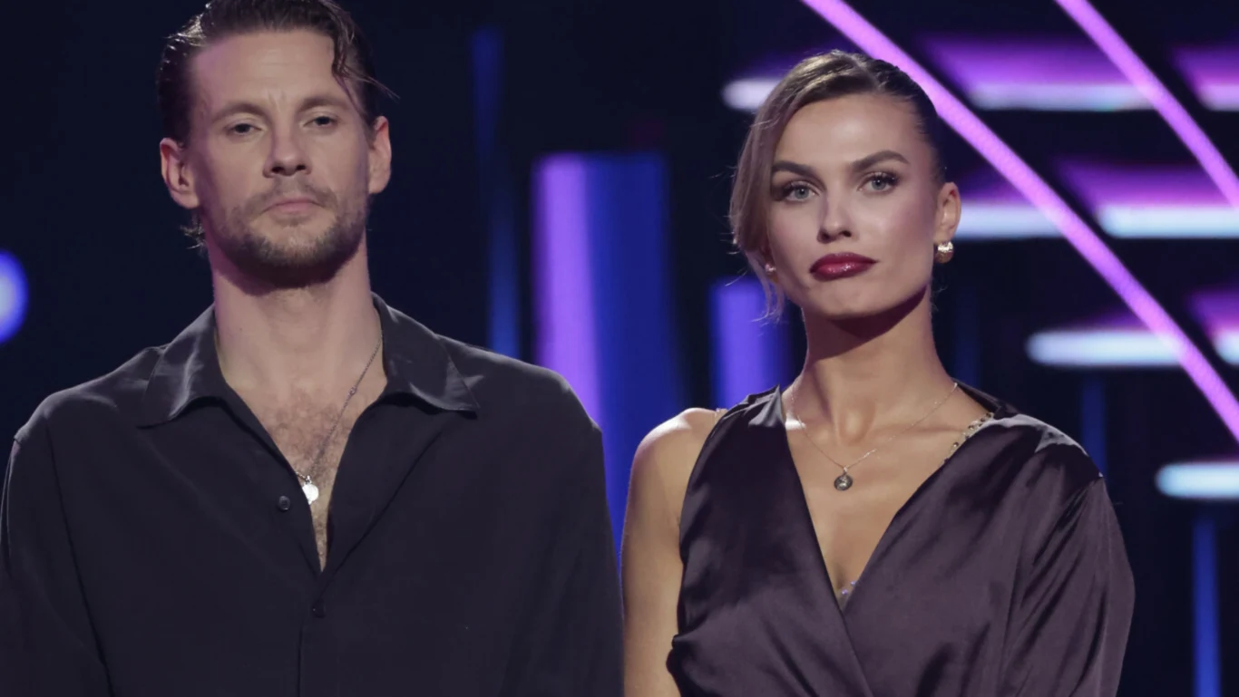 Dwie elegancko ubrane osoby, mężczyzna w ciemnej koszuli i kobieta w wieczorowej sukni, stojące obok siebie na tle oświetlonej sceny z fioletowymi akcentami świetlnymi.