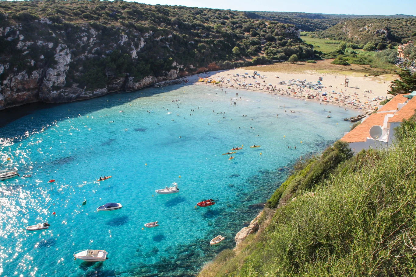 Turkusowa zatoka otoczona zielonymi wzgórzami, piaszczysta plaża pełna wypoczywających osób, liczne łódki i kajaki na spokojnej wodzie oraz niewielkie budynki z czerwonymi dachami w prawym dolnym rogu.