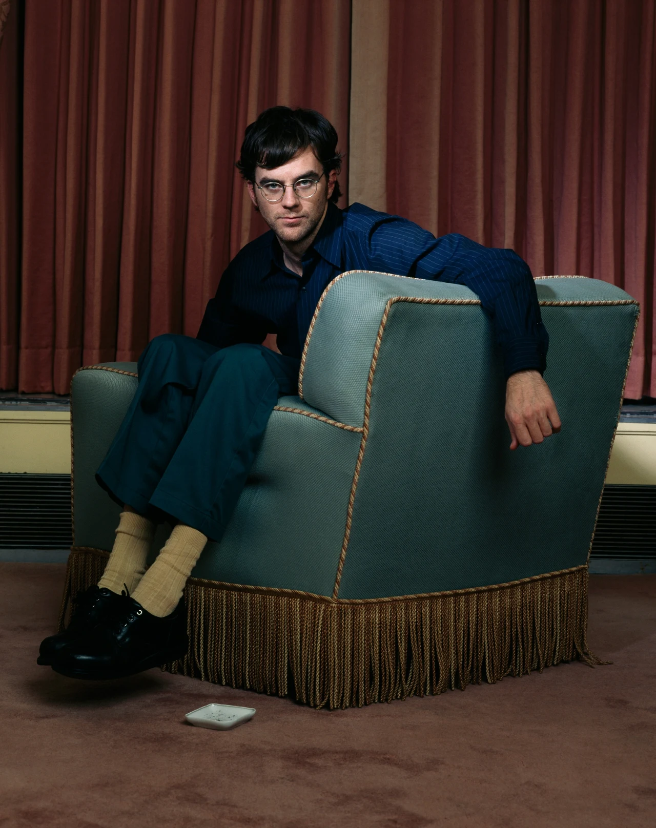 Mężczyzna w okularach siedzi bokiem w dużym, tapicerowanym fotelu z ozdobnymi frędzlami; u jego stóp na dywanie leży biały przedmiot, a tło stanowią ciężkie, zasłonięte zasłony w ciemniejszym odcieniu.