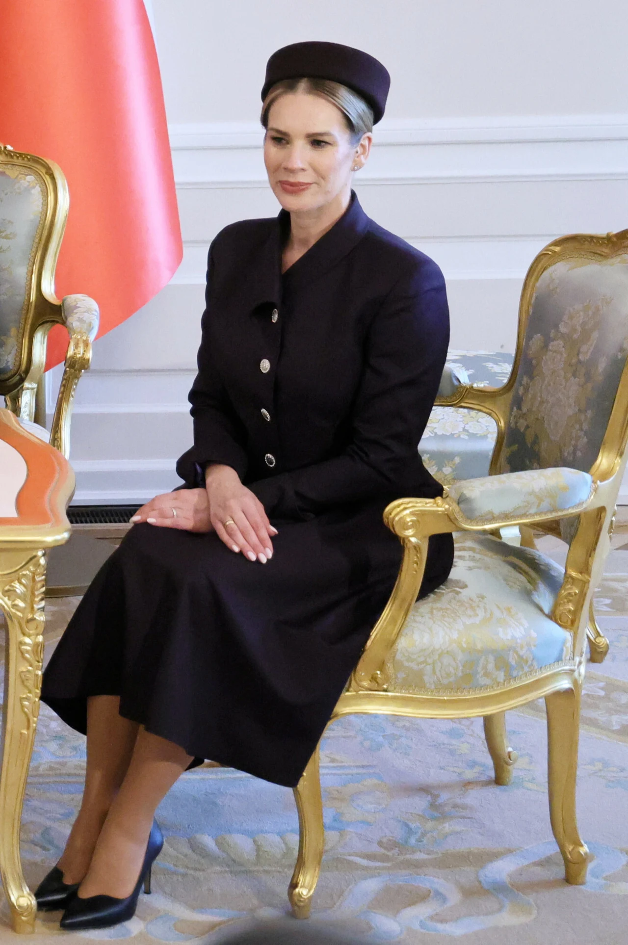 Kobieta w eleganckim, czarnym stroju i kapeluszu siedzi na złotym, ozdobnym krześle, złożone dłonie spoczywają na kolanach; w tle widoczny fragment czerwonej flagi i biało-złote meble o klasycznym stylu.