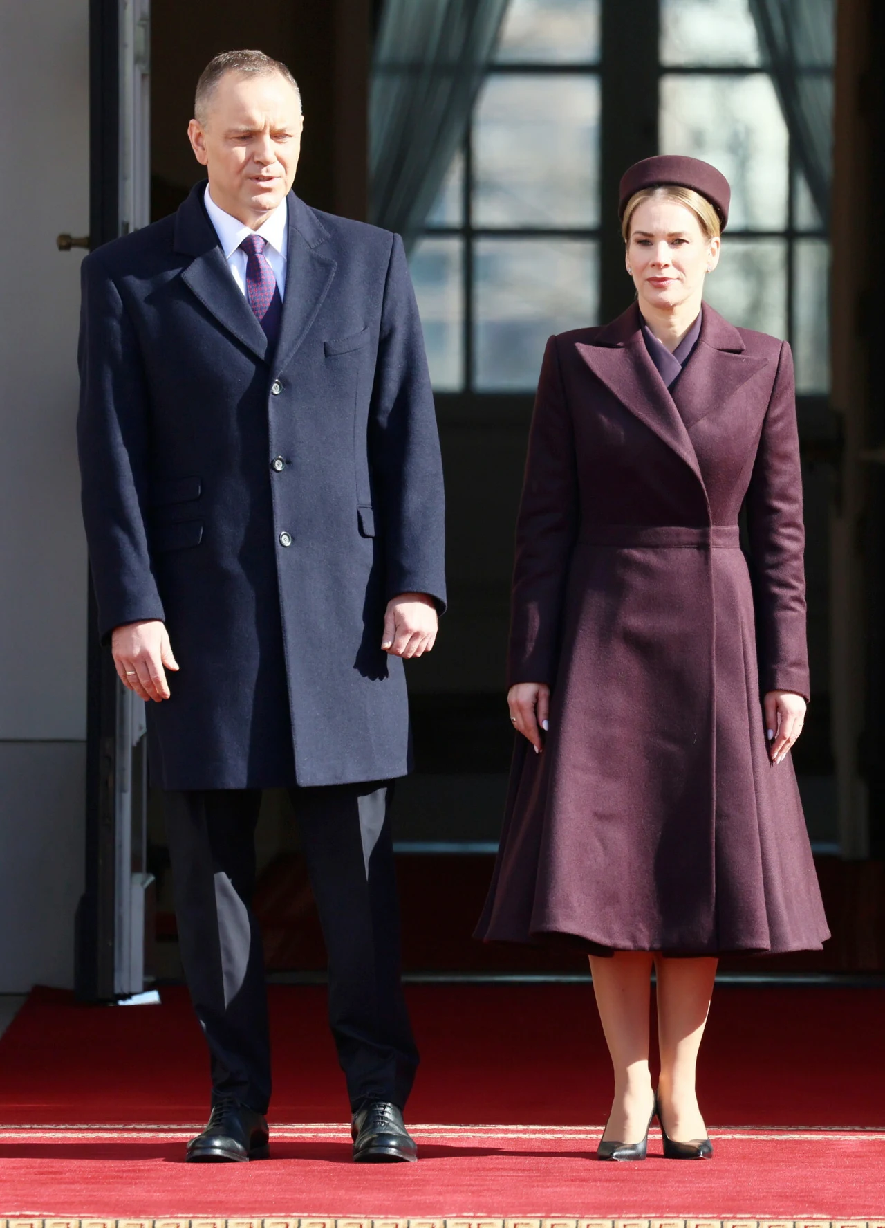 Dwójka elegancko ubranych osób stoi na czerwonym dywanie przed wejściem do budynku, mężczyzna w ciemnym płaszczu i kobieta w bordowym płaszczu oraz dopasowanym kapeluszu patrzą przed siebie, światło słoneczne oświetla ich postacie.