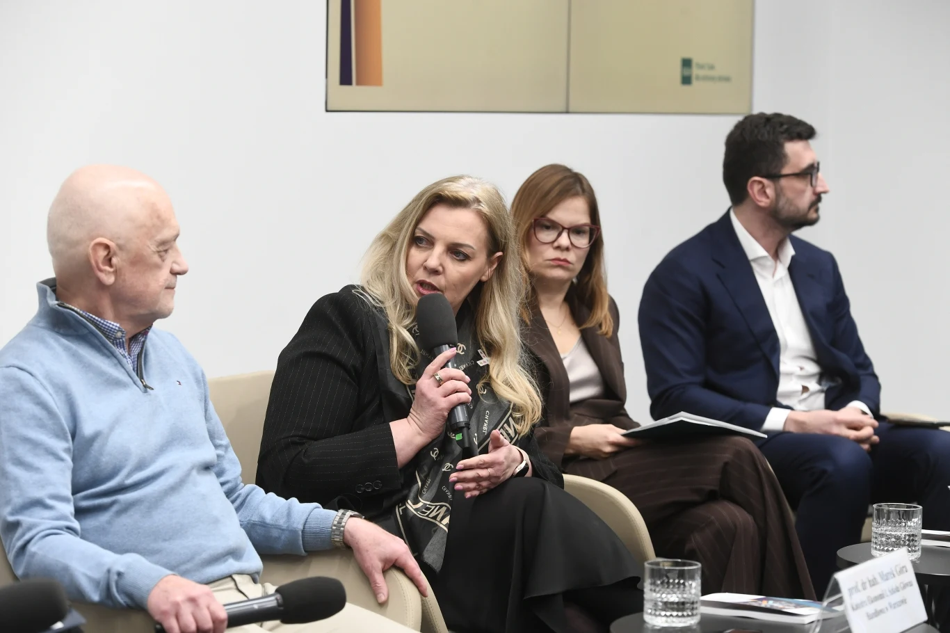 Czworo dorosłych osób siedzi na panelu dyskusyjnym, dwie kobiety i dwóch mężczyzn ujętych w półprofilu; jedna z kobiet trzyma mikrofon i mówi, reszta prezentuje postawę słuchającą lub czeka na swoją kolej do głosu; na stoliku przed nimi stoją szklanki ...