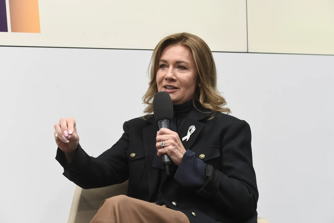 Kobieta z jasnobrązowymi włosami w półdługiej fryzurze siedzi na krześle, trzyma mikrofon i mówi, ubrana w czarną marynarkę z białą wstążką przypiętą do klapy.