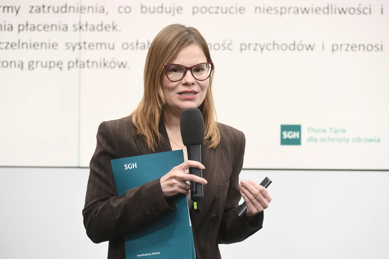 Kobieta w okularach trzyma mikrofon oraz niebieską teczkę z logo SGH, stojąc przed tablicą z tekstem, co wskazuje na prezentację lub wystąpienie eksperckie na tle związanym z tematyką Think Tanku SGH.