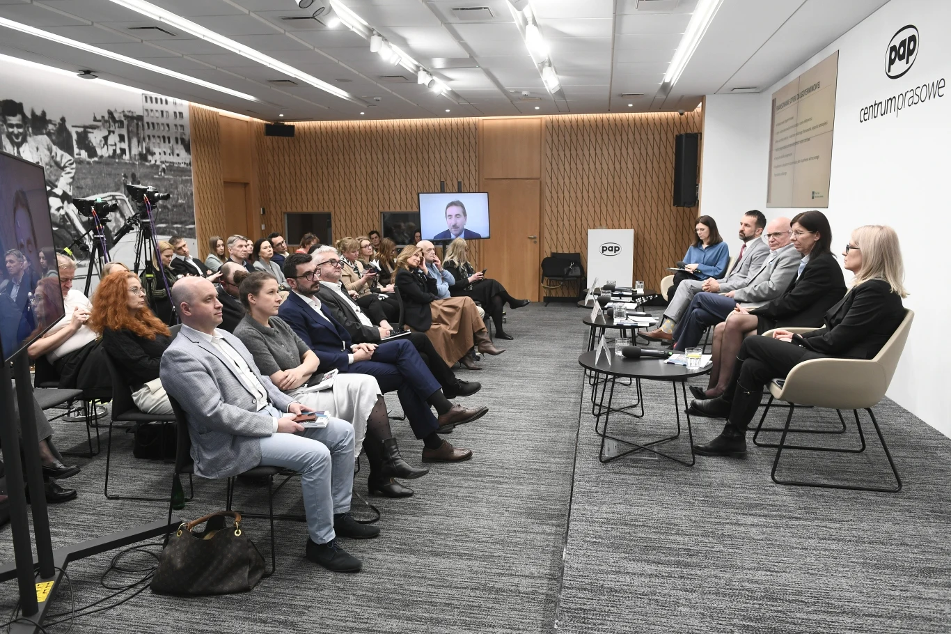 Panel dyskusyjny z udziałem pięciu prelegentów siedzących przy stole, naprzeciwko licznie zgromadzonej publiczności; w tle widoczne ekrany, kamery oraz logo PAP na białej ścianie po prawej stronie.