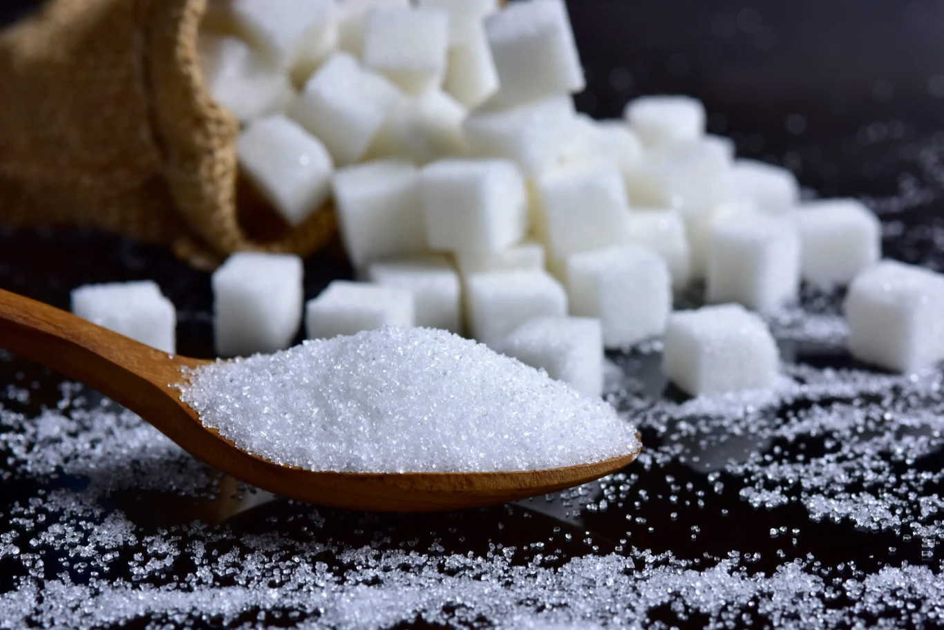 Drewniana łyżka z białym cukrem kryształem na tle rozsypanych kostek cukru oraz fragmentu worka jutowego.