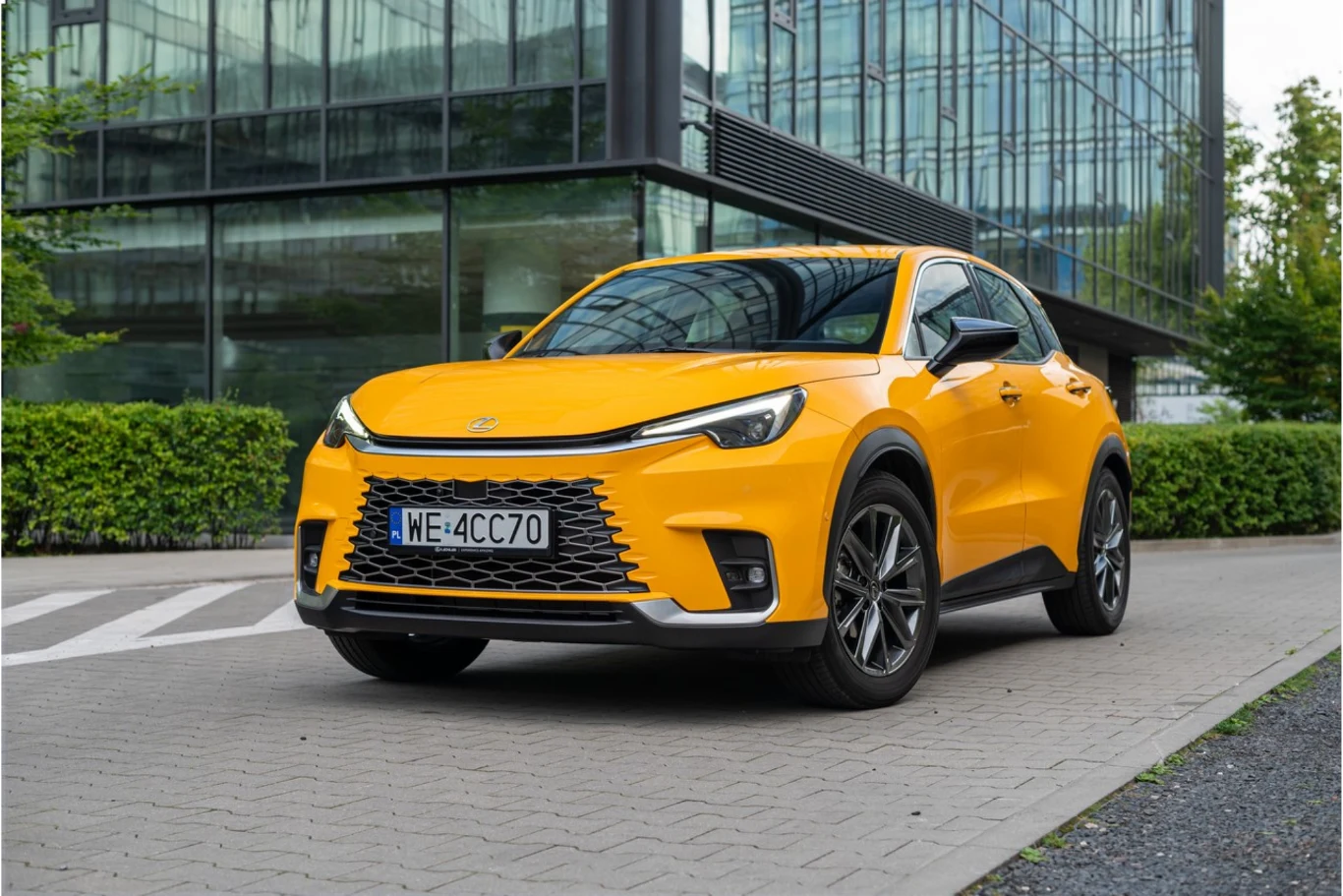 Jakie standardowe wyposażenie znajduje się na liście modelu LBX, UX i NX? Jaskrawożółty SUV Lexus zaparkowany przed przeszklonym biurowcem, z czarnymi felgami i charakterystycznym grillem.