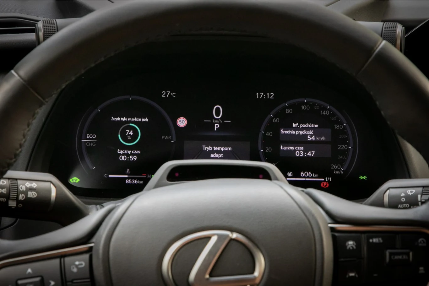 Zestaw wskaźników w modelu UX300h. Cyfrowy zestaw wskaźników z logo Lexusa na kierownicy, wyświetlający aktualne informacje o prędkości, trybie jazdy, zasięgu, zużyciu paliwa oraz temperaturze otoczenia; widoczne także przyciski na kierownicy i elementy kokpitu.