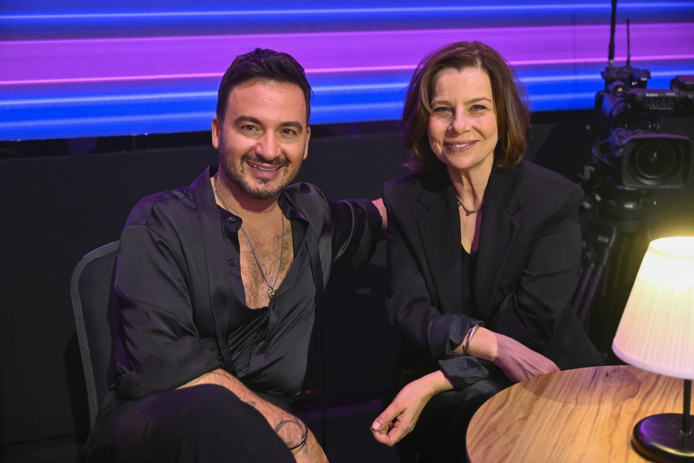 Dwoje uśmiechniętych ludzi siedzi obok siebie przy okrągłym stole, ubrani elegancko na czarno, w tle widoczne są kolorowe światła i kamera studyjna, na pierwszym planie stoi oświetlona lampa stołowa.