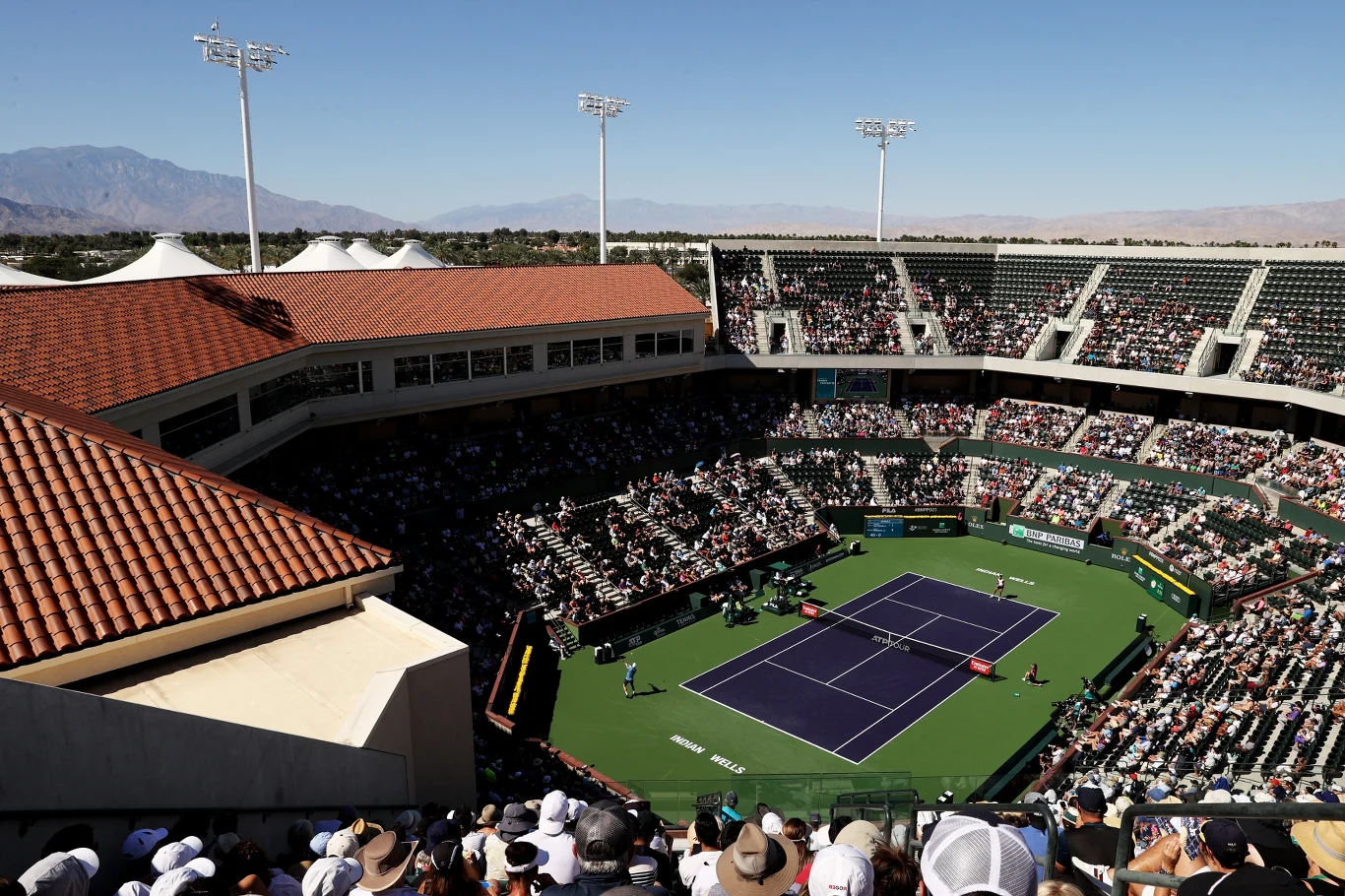 Duży stadion tenisowy otoczony trybunami wypełnionymi kibicami, centralnie na korcie rozgrywany jest mecz tenisowy, widoczne są dwie zawodniczki oraz sędzia, a w tle widać góry i bezchmurne niebo.