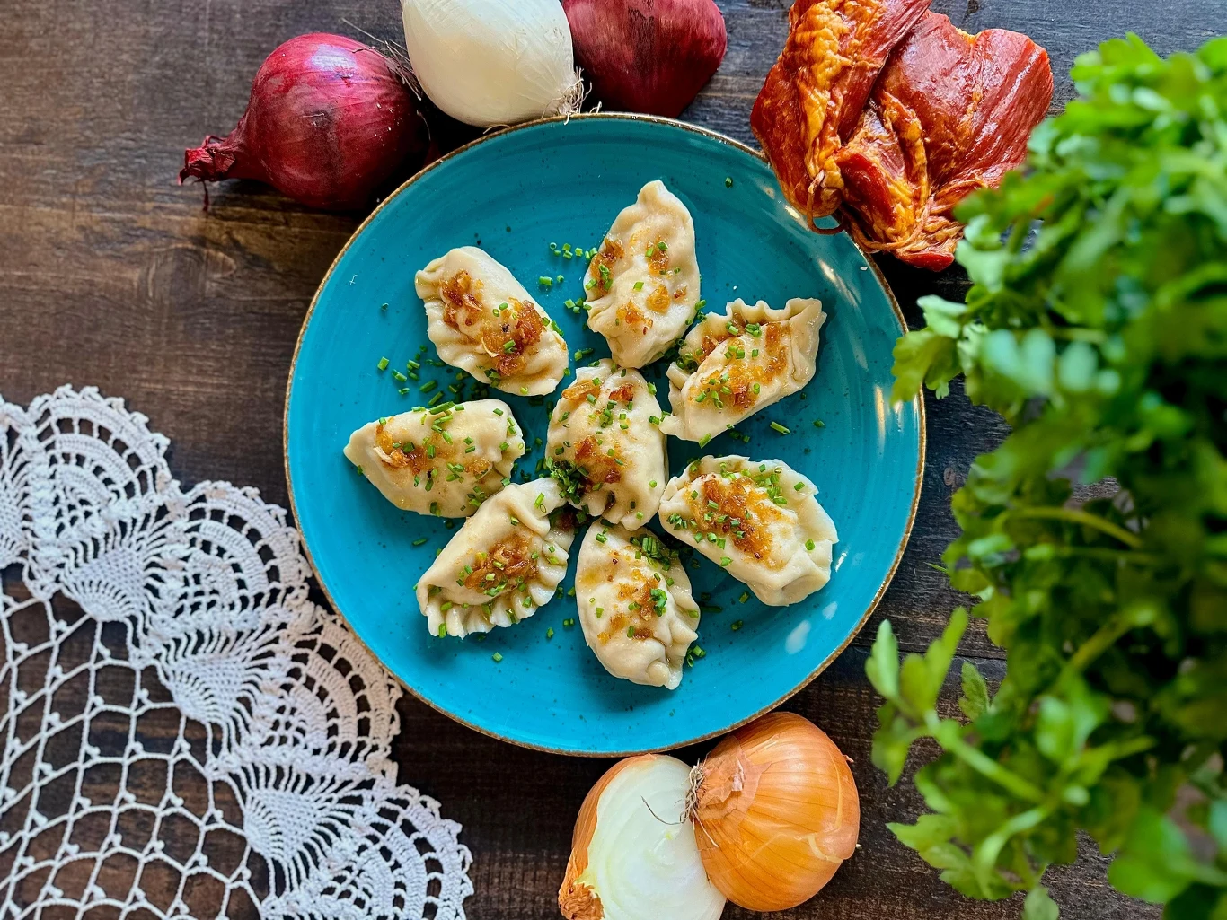 Pierogi udekorowane podsmażoną cebulką na niebieskim talerzu, otoczone świeżą natką pietruszki, cebulami i suszoną papryką, na ciemnobrązowym drewnianym stole z fragmentem białej serwetki koronki.