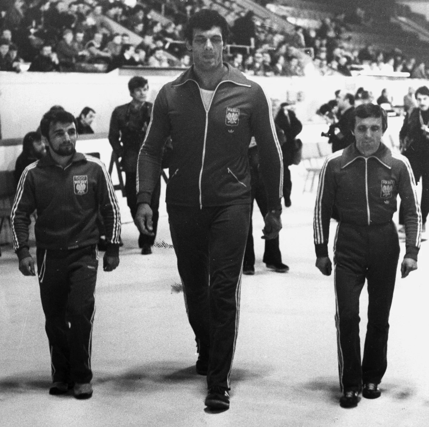 Trzech sportowców w dresach z orłem na piersi idzie obok siebie po hali sportowej, wyraźnie wyróżniając się wzrostem – środkowy mężczyzna jest znacznie wyższy od pozostałych dwóch.