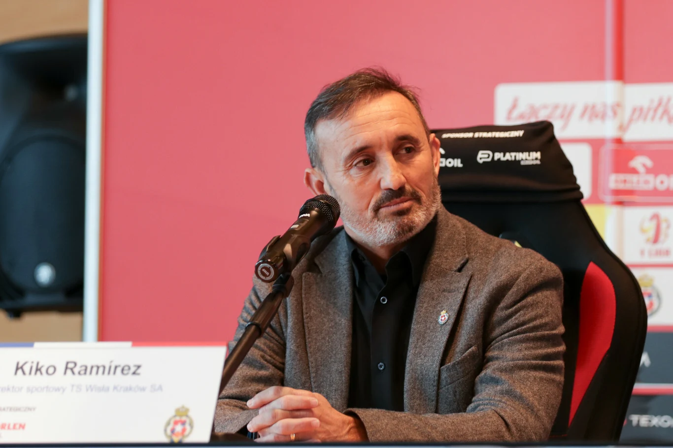 Mężczyzna w eleganckiej marynarce siedzi przy stole konferencyjnym, trzyma przed sobą mikrofon, w tle widoczna czerwona ścianka z logotypami i napisami związanymi z piłką nożną.