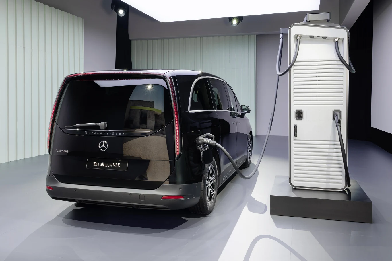 Czarny elektryczny van marki Mercedes-Benz podłączony do stacji ładowania w nowoczesnym pomieszczeniu o jasnym oświetleniu i minimalistycznym wystroju.