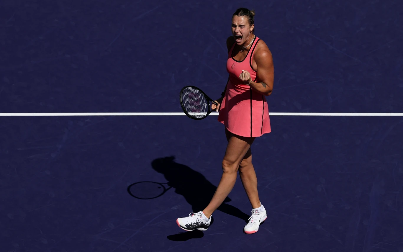 Tenisistka w różowej sukience energicznie zaciska pięść i wyraża silne emocje po udanym punkcie na korcie tenisowym, trzymając rakietę w lewej ręce.