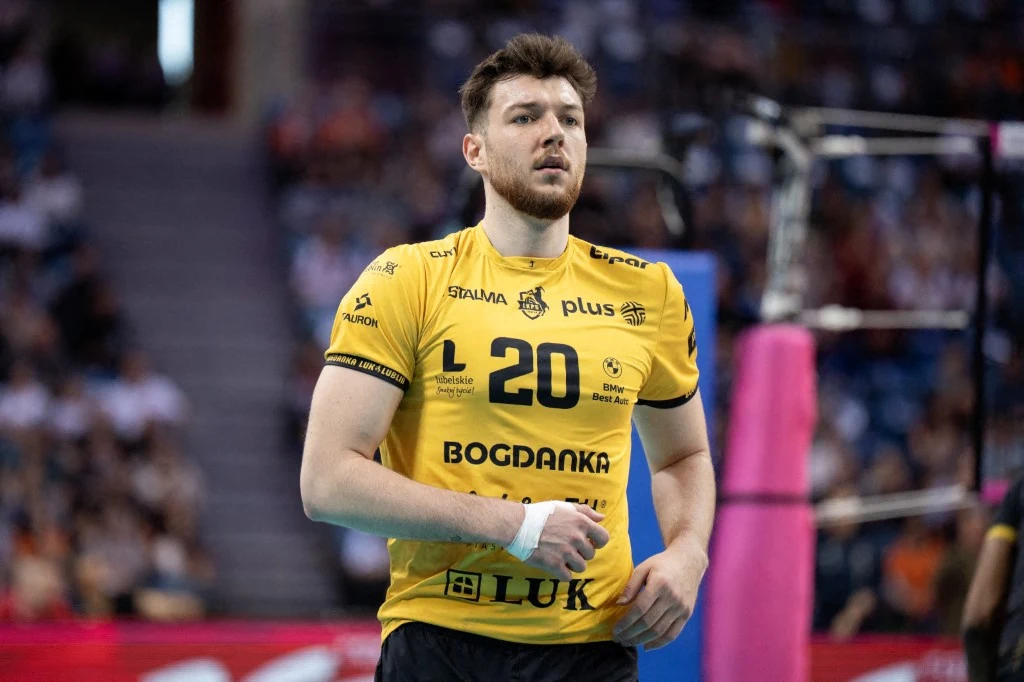 Siatkarz w żółtej koszulce z numerem 20, oznaczeniami sponsorskimi i nazwą drużyny, stoi na boisku podczas meczu, w tle widoczne są elementy siatki oraz rozmyte sylwetki publiczności na trybunach.