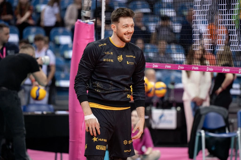 Siatkarz w czarnym stroju sportowym stoi uśmiechnięty obok siatki na hali sportowej podczas rozgrywek, w tle widoczna jest piłka do siatkówki oraz kibice na trybunach.