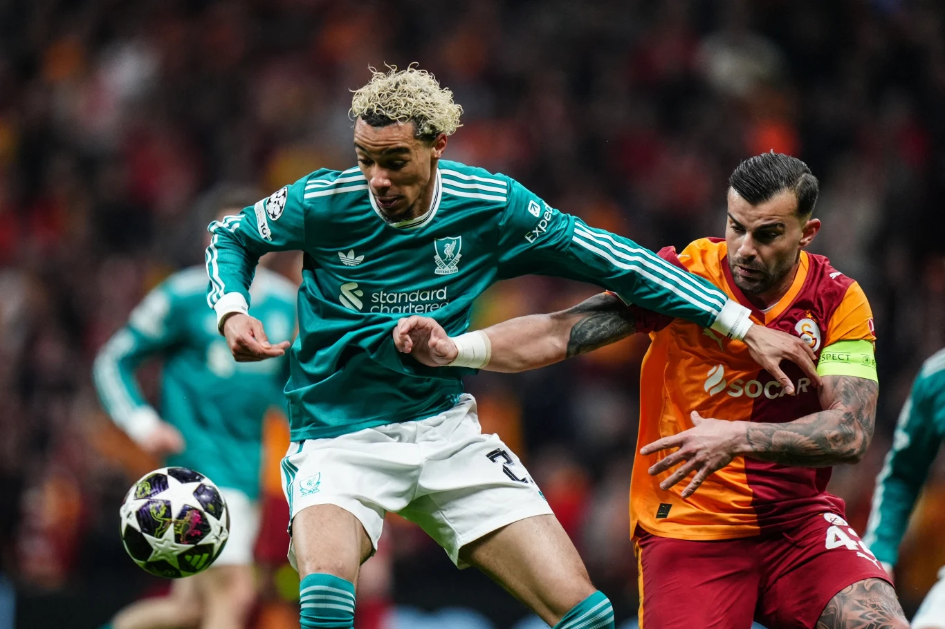 Dwóch piłkarzy walczy o piłkę podczas meczu; zawodnik w zielonej koszulce blokowany jest przez rywala w pomarańczowo-czerwonym stroju, obaj prezentują intensywną rywalizację fizyczną.