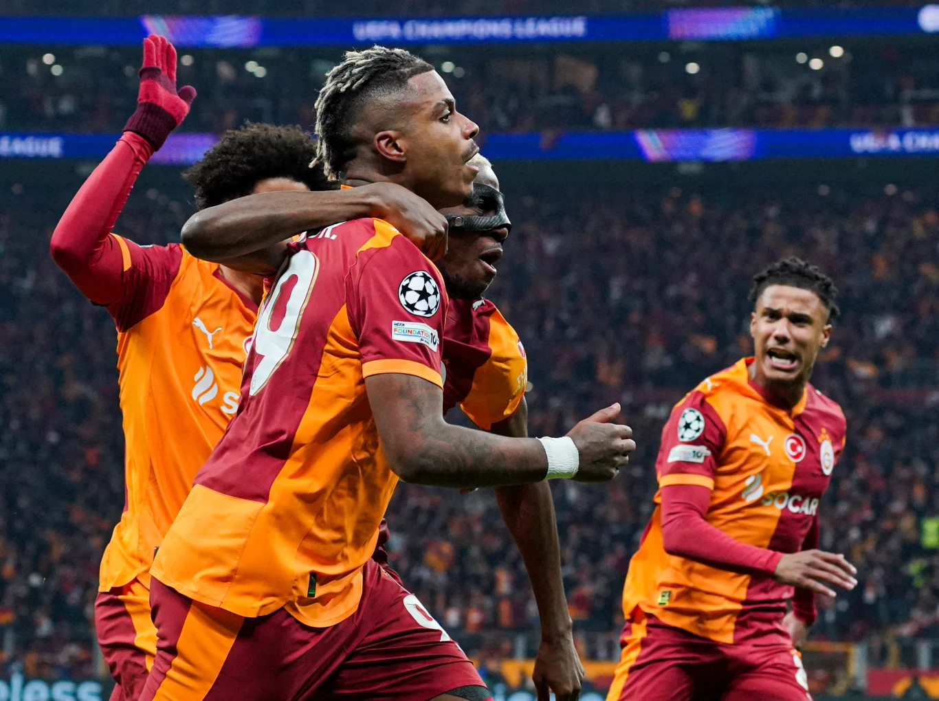 Piłkarze Galatasaray w pomarańczowo-czerwonych strojach świętują zdobycie bramki podczas meczu Ligi Mistrzów, wyrażając radość i emocje na tle pełnego kibiców stadionu.