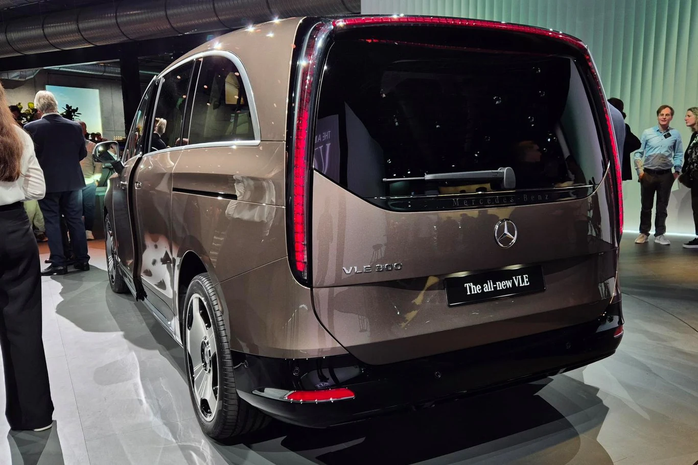 Brązowy elektryczny minivan Mercedes-Benz VLE 300 z wyraźnym logo marki, dużym tylnym oknem i nowoczesnym, aerodynamicznym designem, prezentowany w oświetlonym wnętrzu podczas wydarzenia motoryzacyjnego; w tle kilka osób obserwujących pojazd.