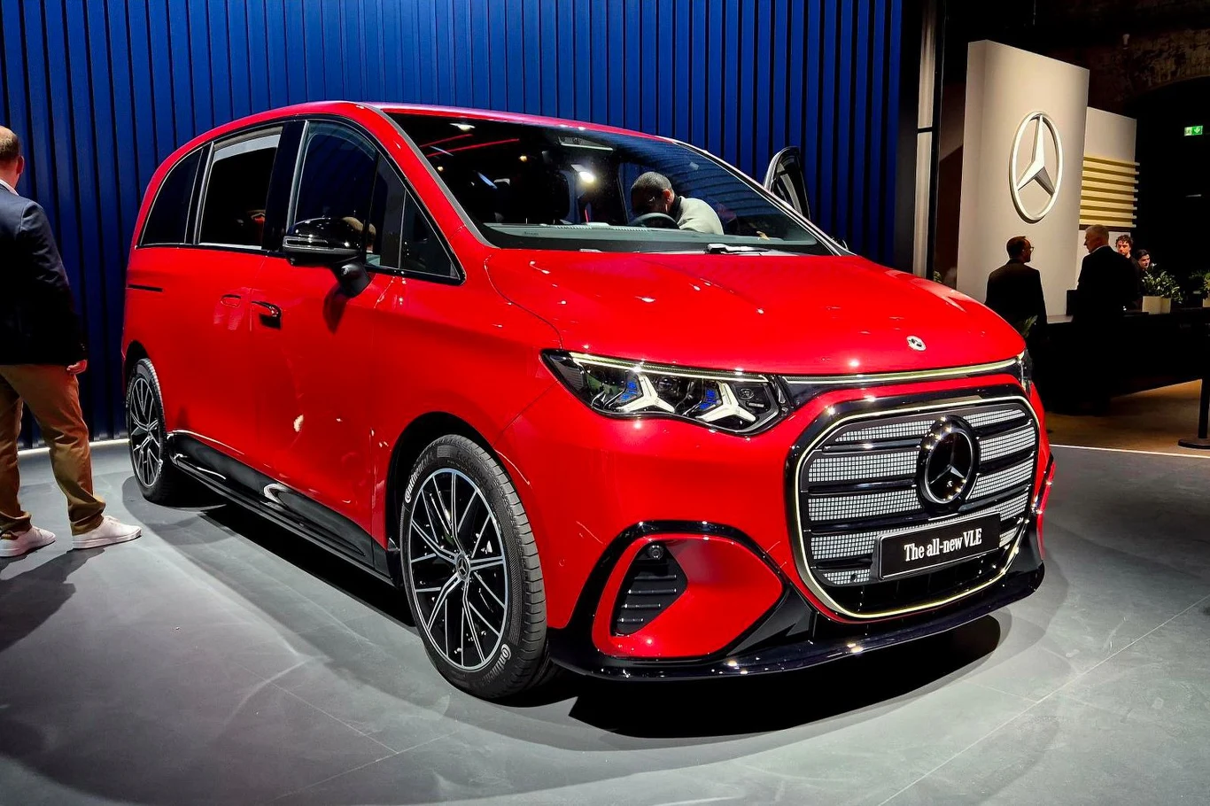 Czerwony minivan Mercedes z charakterystycznym osłoną chłodnicy, ciemnymi szybami i nowoczesnym oświetleniem LED ustawiony w przestrzeni wystawowej na tle niebieskiej ścianki z logo marki.
