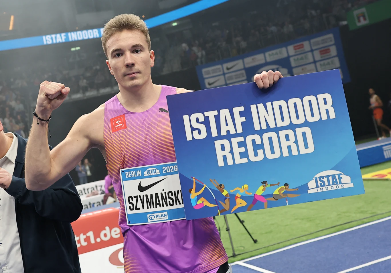 Lekkoatleta w sportowym stroju unosi zaciśniętą pięść w geście triumfu, trzymając tabliczkę z napisem 'ISTAF INDOOR RECORD', na arenie lekkoatletycznej z widoczną publicznością w tle.