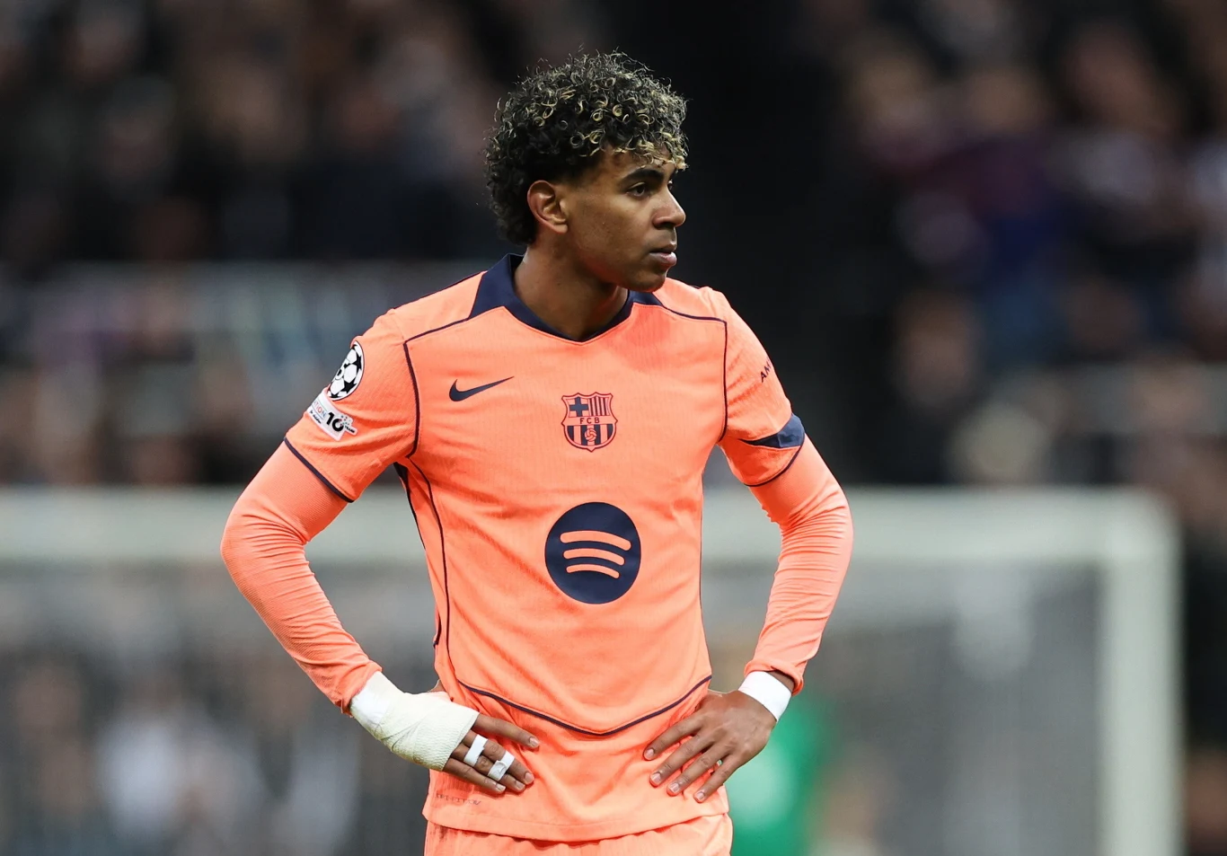 Piłkarz w pomarańczowym stroju FC Barcelony z opaską na nadgarstku stoi na boisku z rękami na biodrach, skoncentrowany i skierowany w bok, w tle rozmazana publiczność stadionu.
