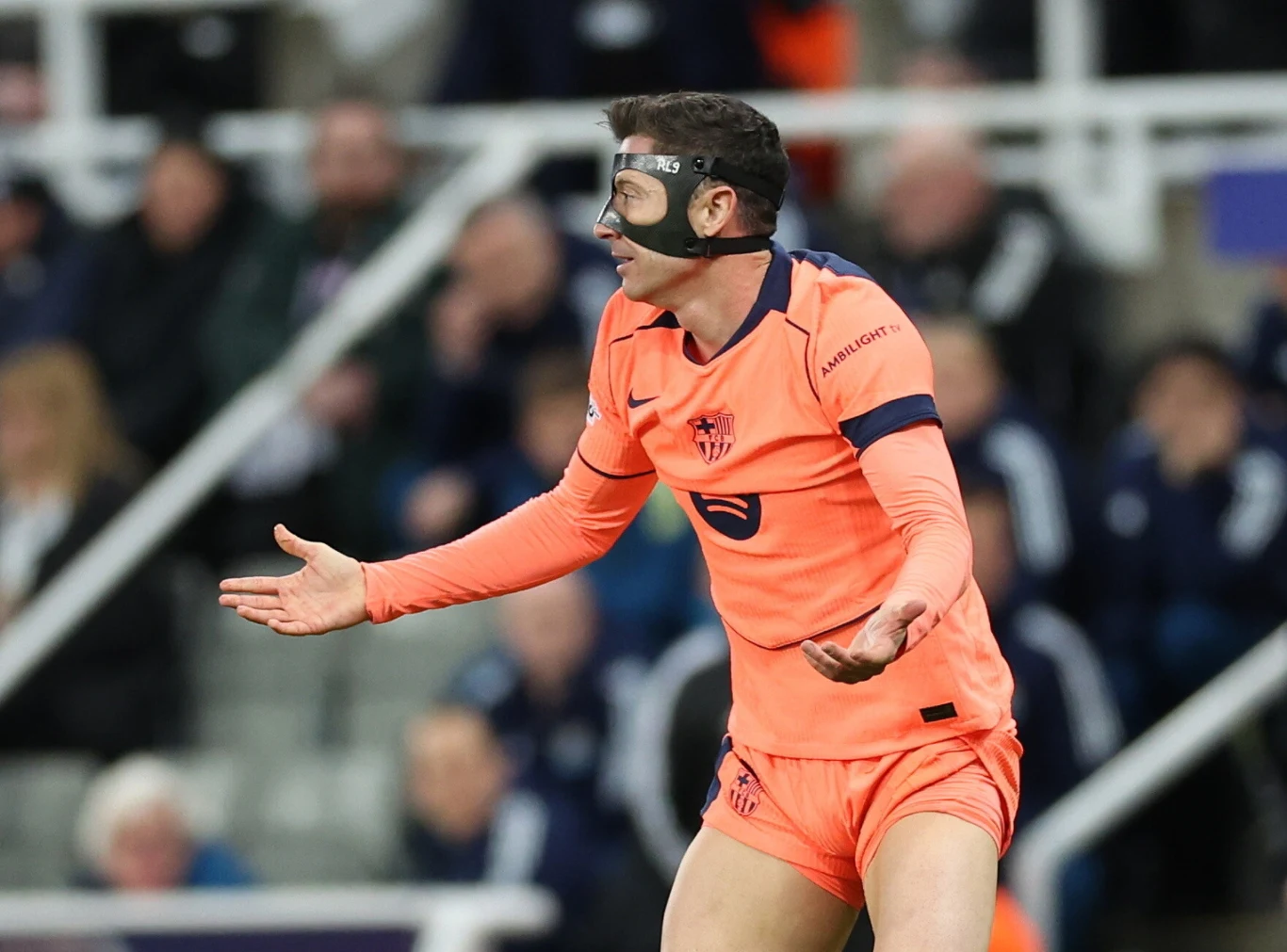 Piłkarz w pomarańczowym stroju z czarną maską ochronną na twarzy gestykuluje z rozłożonymi rękami na boisku podczas meczu, w tle rozmyta publiczność na trybunach.