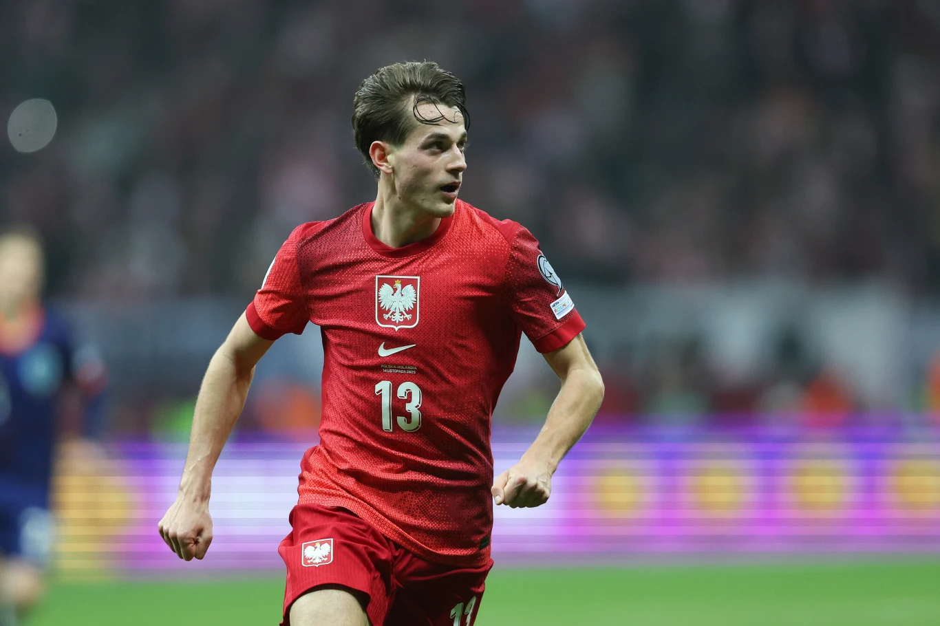 Piłkarz reprezentacji Polski w czerwonym stroju z numerem 13 na koszulce biegnie po murawie stadionu podczas meczu, w tle rozmazana publiczność i elementy oświetlenia stadionowego.