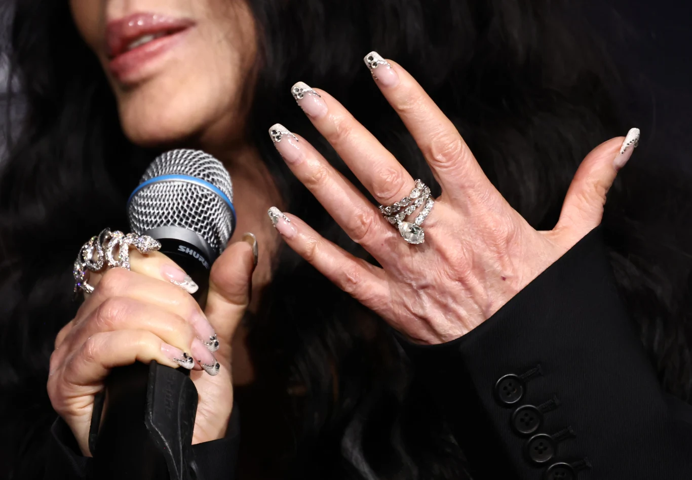 Kobieta z długimi, ozdobionymi paznokciami i licznymi srebrnymi pierścionkami trzyma mikrofon, eksponując dłoń oraz biżuterię na palcach, usta delikatnie zarysowane szminką.