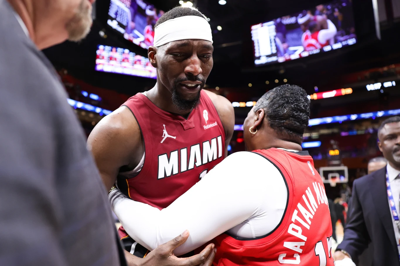 Koszykarz drużyny Miami w bordowym stroju obejmuje osobę w czerwonej kamizelce z napisem 'CAPTAIN' na plecach, otoczony przez innych ludzi na parkiecie hali sportowej po zakończonym meczu.