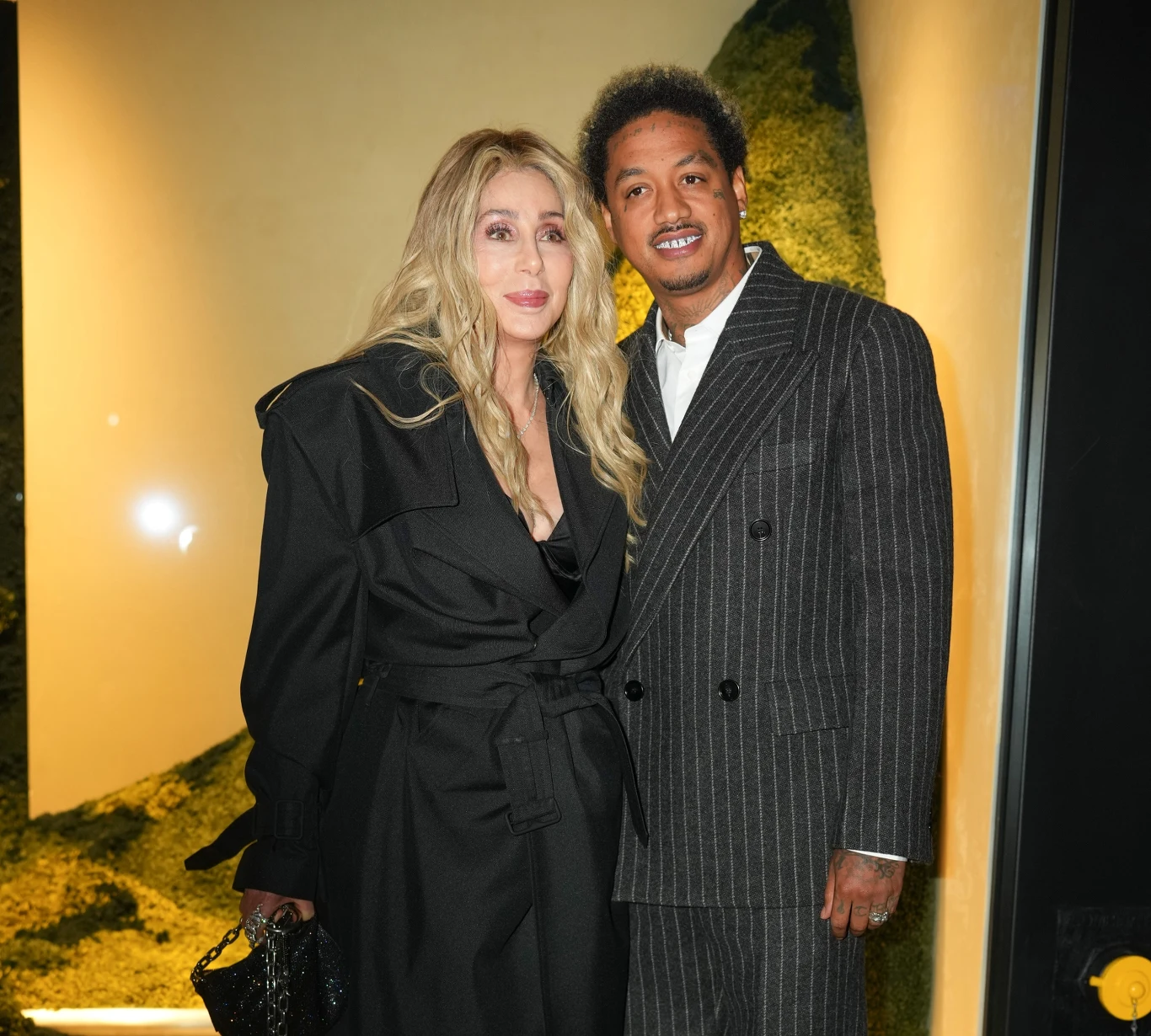 Kobieta w czarnym płaszczu z długimi blond włosami stoi obok mężczyzny w ciemnym, prążkowanym garniturze, oboje uśmiechają się i pozują razem na tle zielonej ściany z roślinnością.