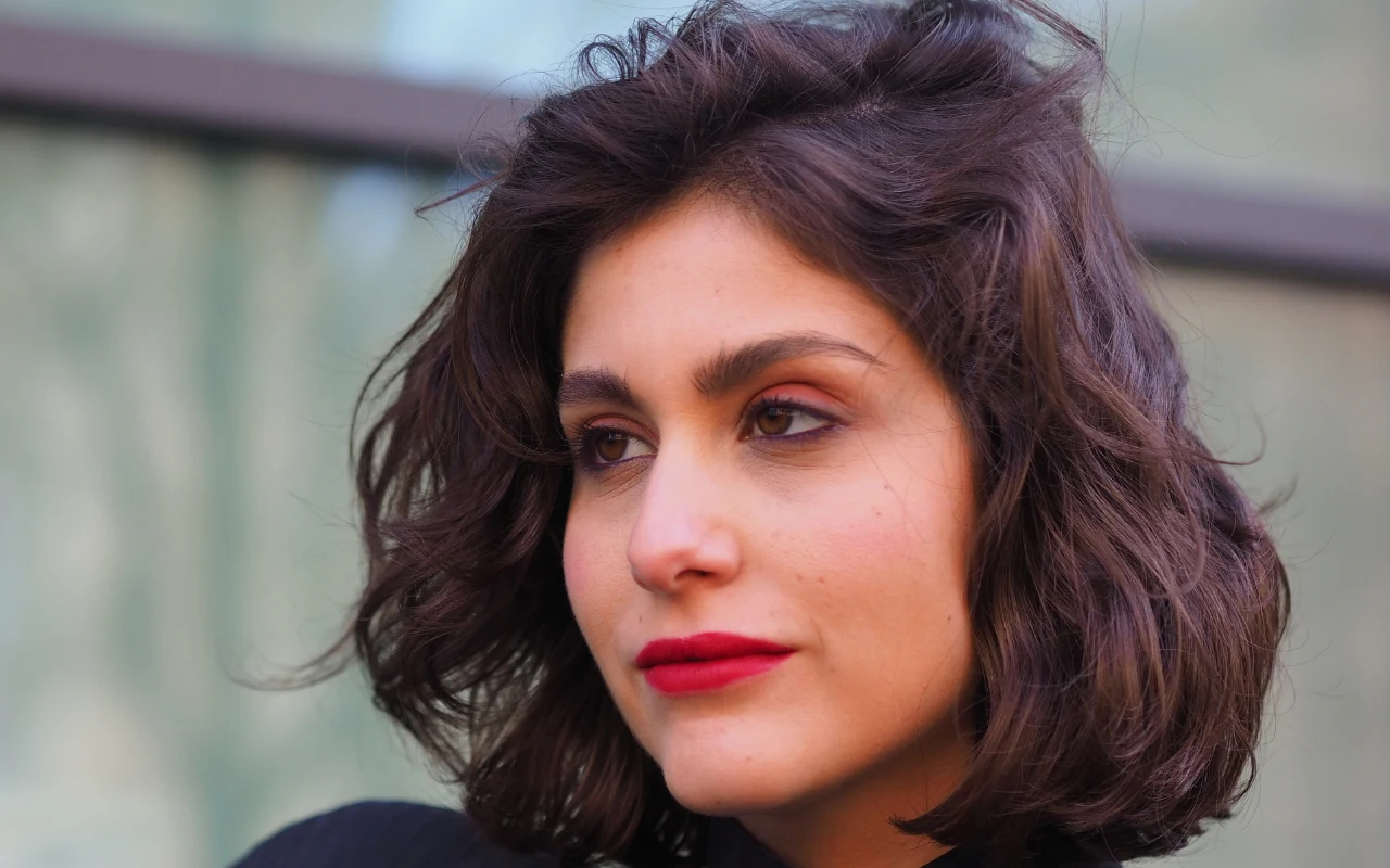 Kobieta o krótkich, ciemnobrązowych włosach i wyrazistym makijażu z czerwonymi ustami, patrząca w bok na neutralnym tle.