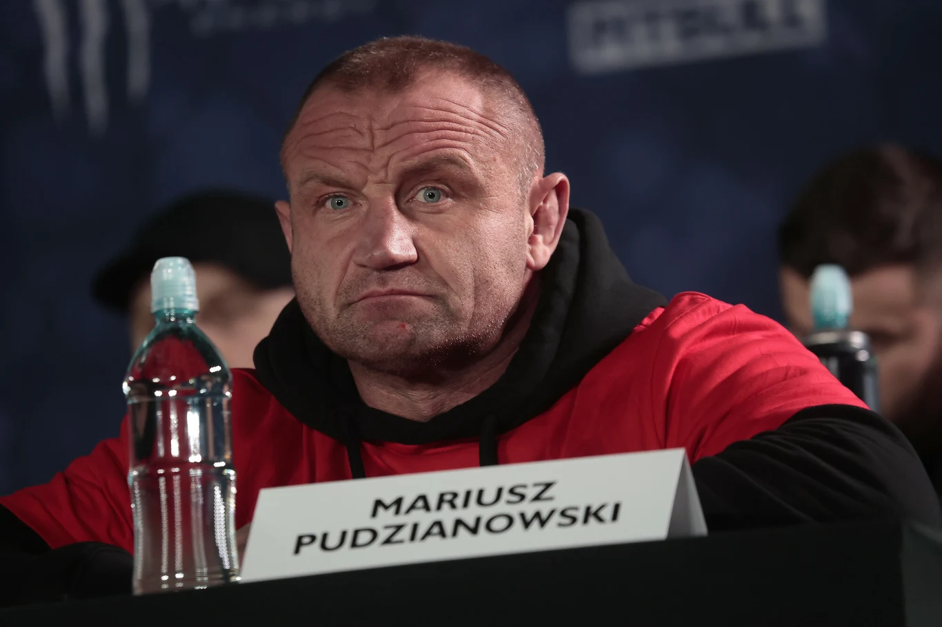 Mężczyzna w czerwonej koszulce siedzi przy stole z tabliczką z napisem „Mariusz Pudzianowski”, obok znajduje się butelka wody, a w tle widoczna jest ciemna sceneria konferencyjna.