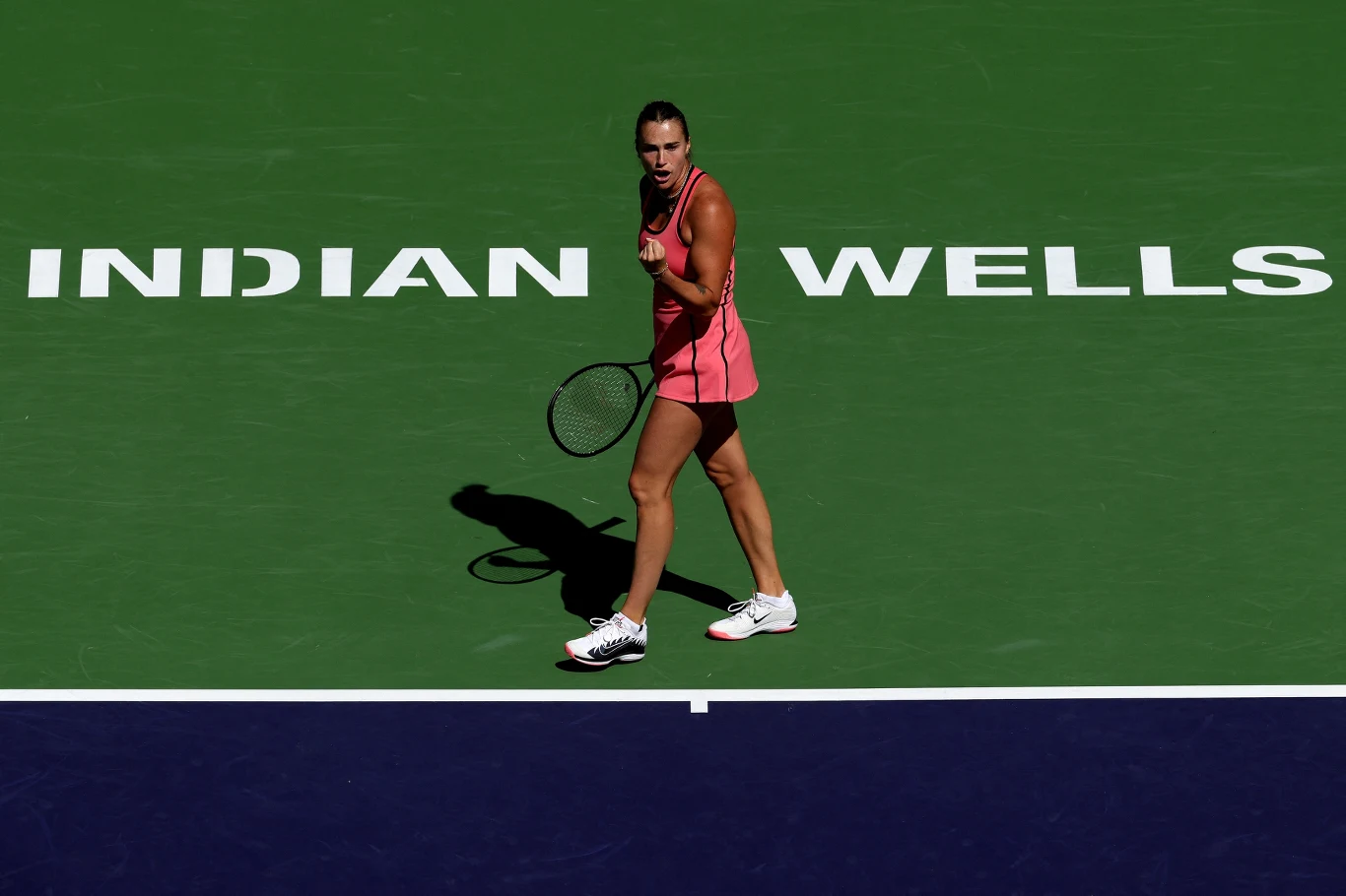 Tenisistka w różowej sukience stoi na korcie z rakietą w ręku, zaciśnięta pięść wyraża silne emocje; w tle duży napis 'INDIAN WELLS' na zielonej nawierzchni kortu.