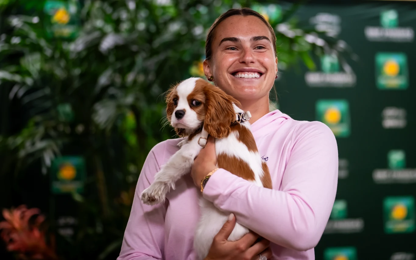 Uśmiechnięta kobieta w różowej bluzie trzyma na rękach małego psa rasy cavalier king charles spaniel, w tle zielone rośliny oraz tablice z logo turnieju tenisowego.