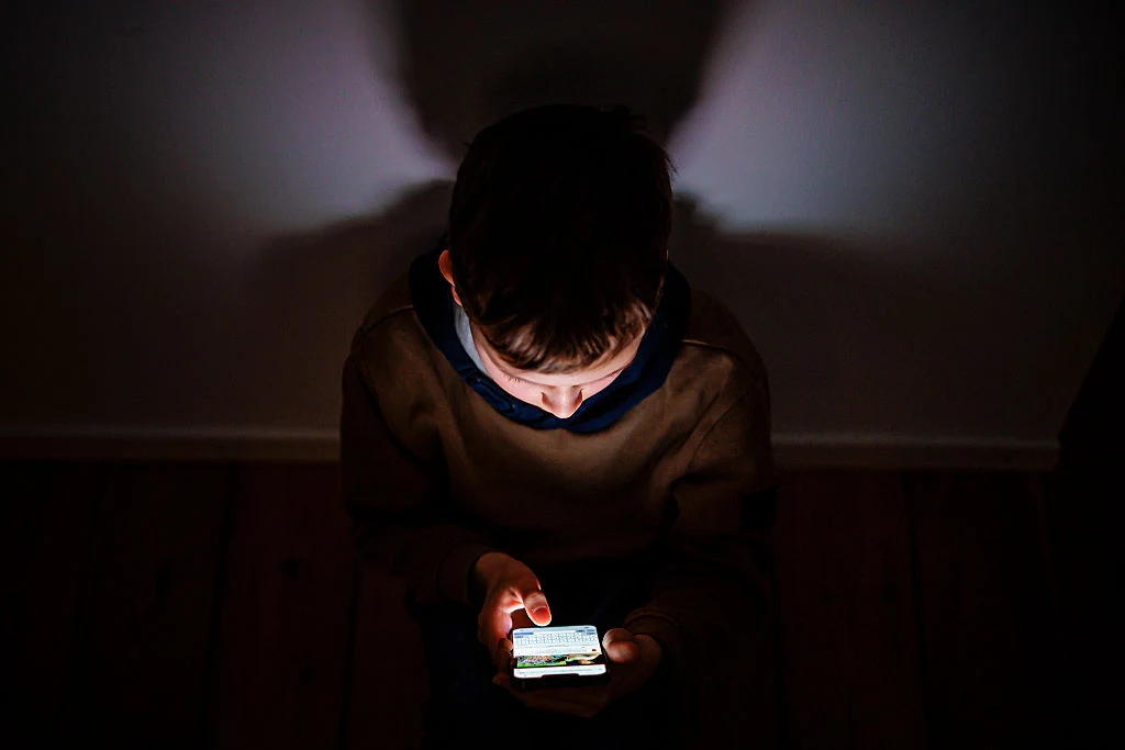 Chłopiec siedzący w ciemnym pomieszczeniu, rozświetlony światłem ekranu smartfona, skupiony na urządzeniu w swoich dłoniach.