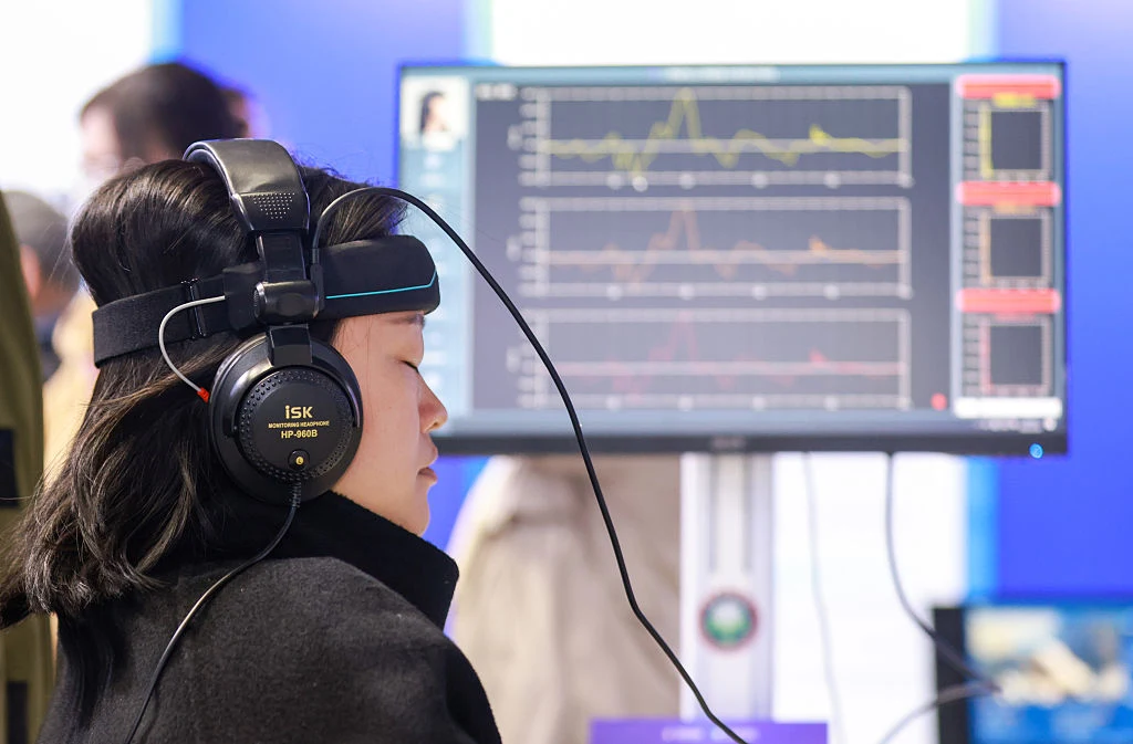 Kobieta z założonym specjalistycznym zestawem słuchawkowym i opaską do pomiaru fal mózgowych siedzi przed monitorem, który wyświetla wykresy danych biometrycznych lub aktywności mózgu.