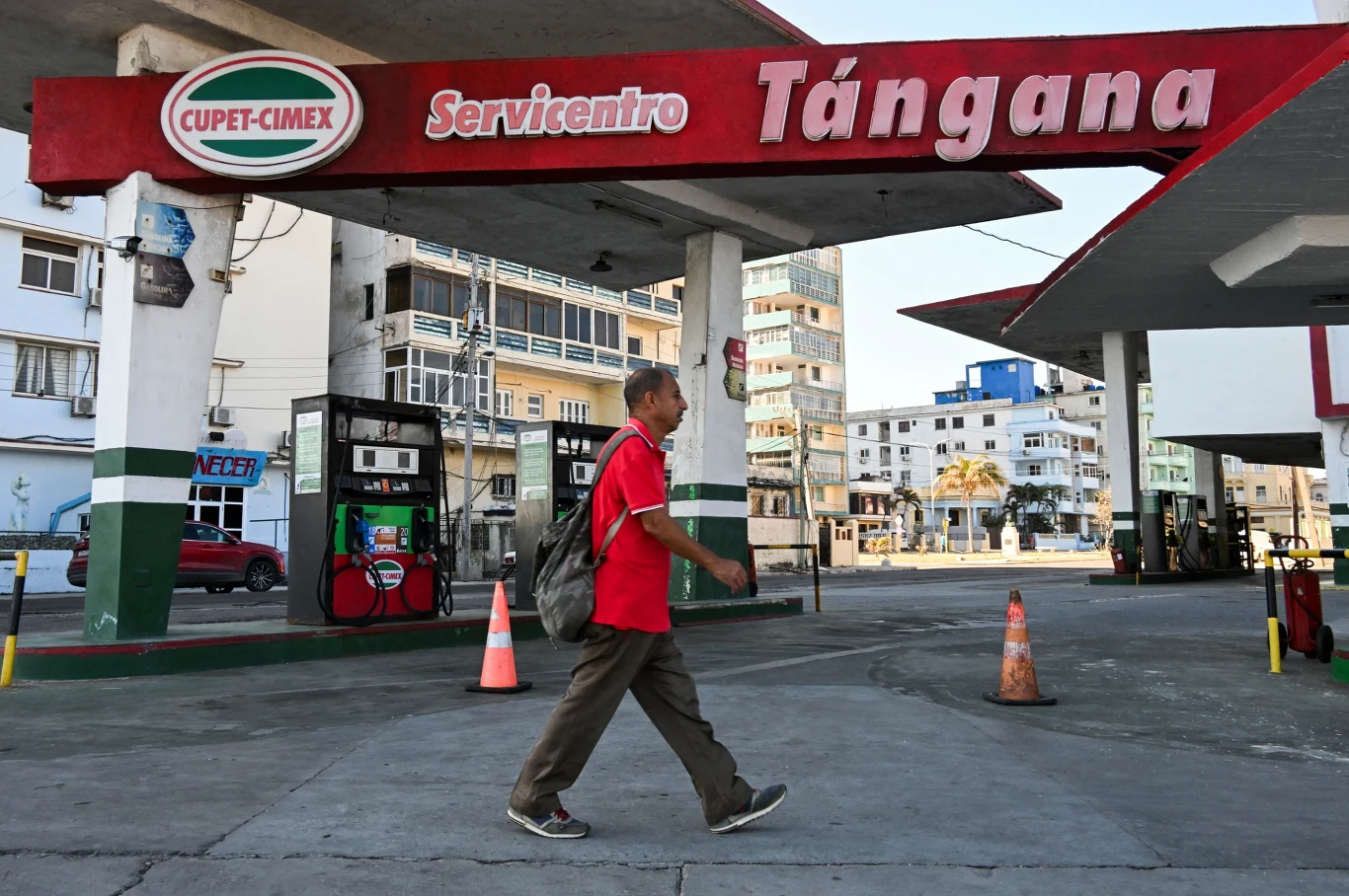 Mężczyzna w czerwonej koszuli i z plecakiem przechodzi przez opustoszałą stację benzynową z widocznym szyldem 'Tángana', w tle nowoczesne budynki miejskie i pachołki drogowe blokujące dostęp do dystrybutorów.