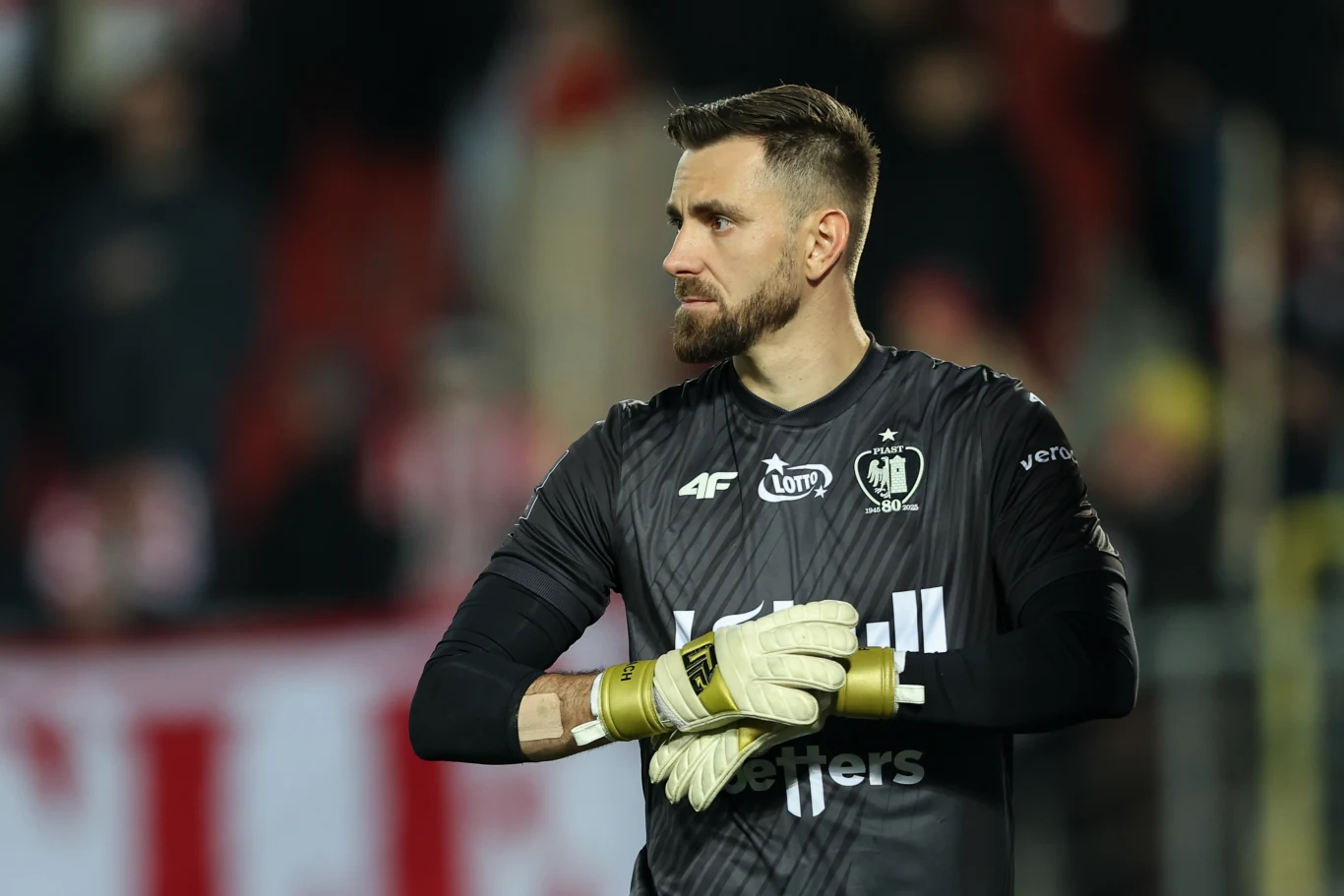 Bramkarz w czarnym stroju sportowym z założonymi rękoma i rękawicami bramkarskimi stoi na boisku podczas meczu, w tle rozmyta publiczność na trybunach.