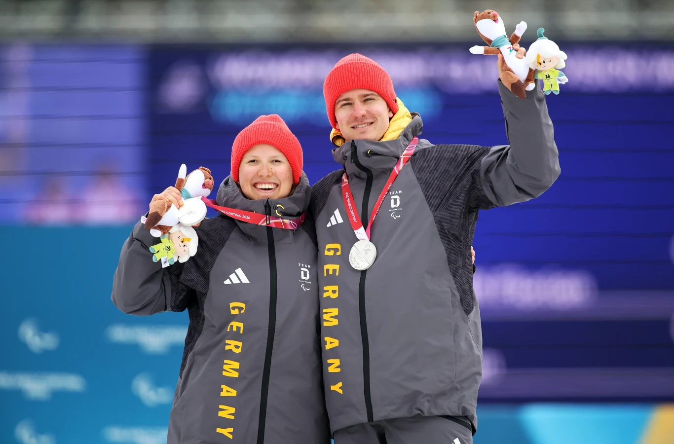Dwoje sportowców z Niemiec w szarych kurtkach z żółtymi napisami i czerwonych czapkach, uśmiechnięci, trzymają srebrne medale oraz maskotki zwycięzcy, stojąc razem na tle niebieskiego ekranu.