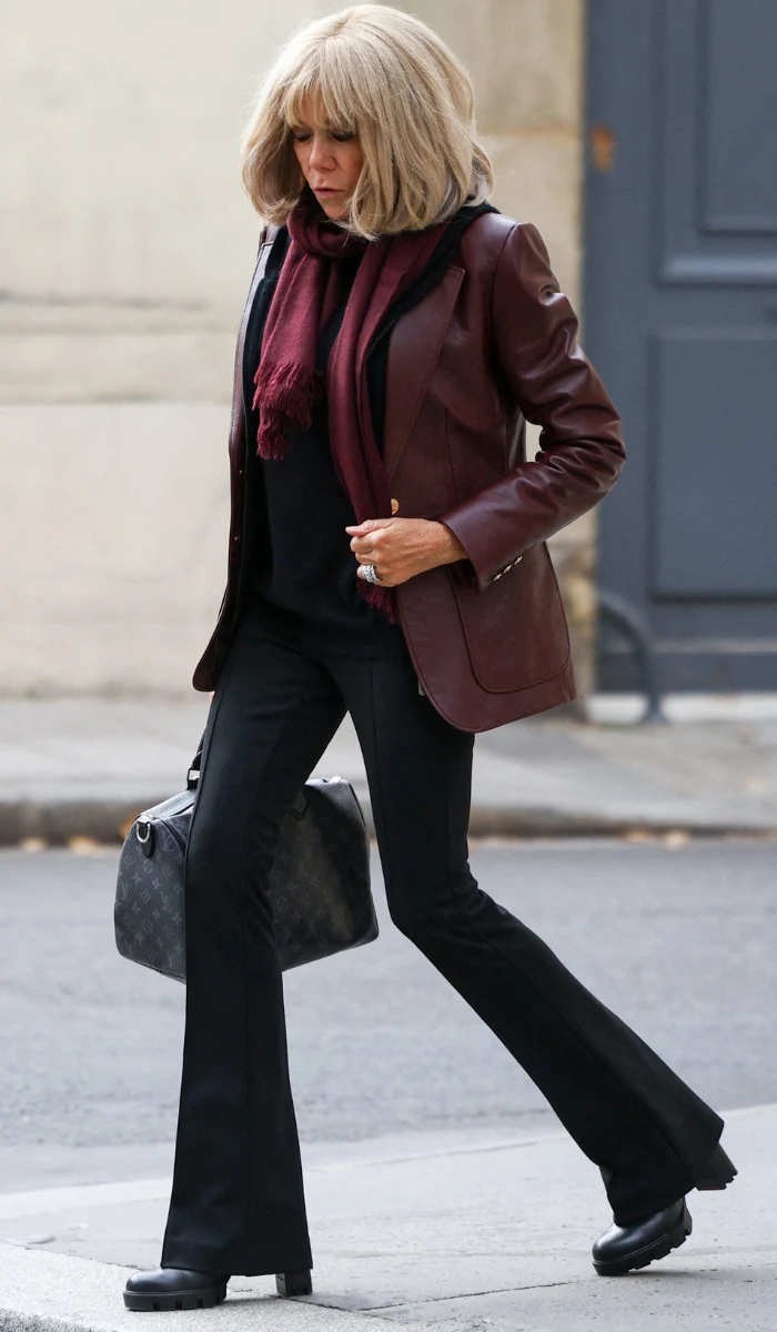 Kobieta z jasnymi włosami ubrana w bordową skórzaną marynarkę, czarny top i czarne spodnie dzwony, z bordowym szalikiem na szyi oraz czarną torebką w ręku, idąca chodnikiem miejskim.