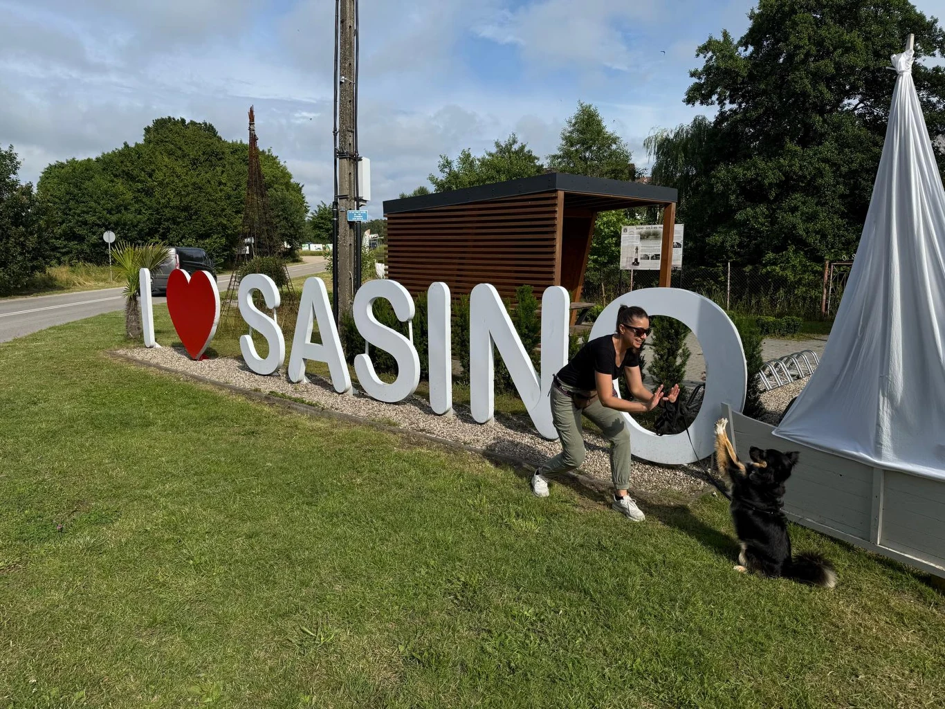 Duże białe litery tworzące napis 'I ♥ SASINO' na tle zielonego trawnika, obok którego kobieta przyklęka i podaje łapę czarnemu psu, w tle widoczne drzewa oraz nowoczesna drewniana altana.