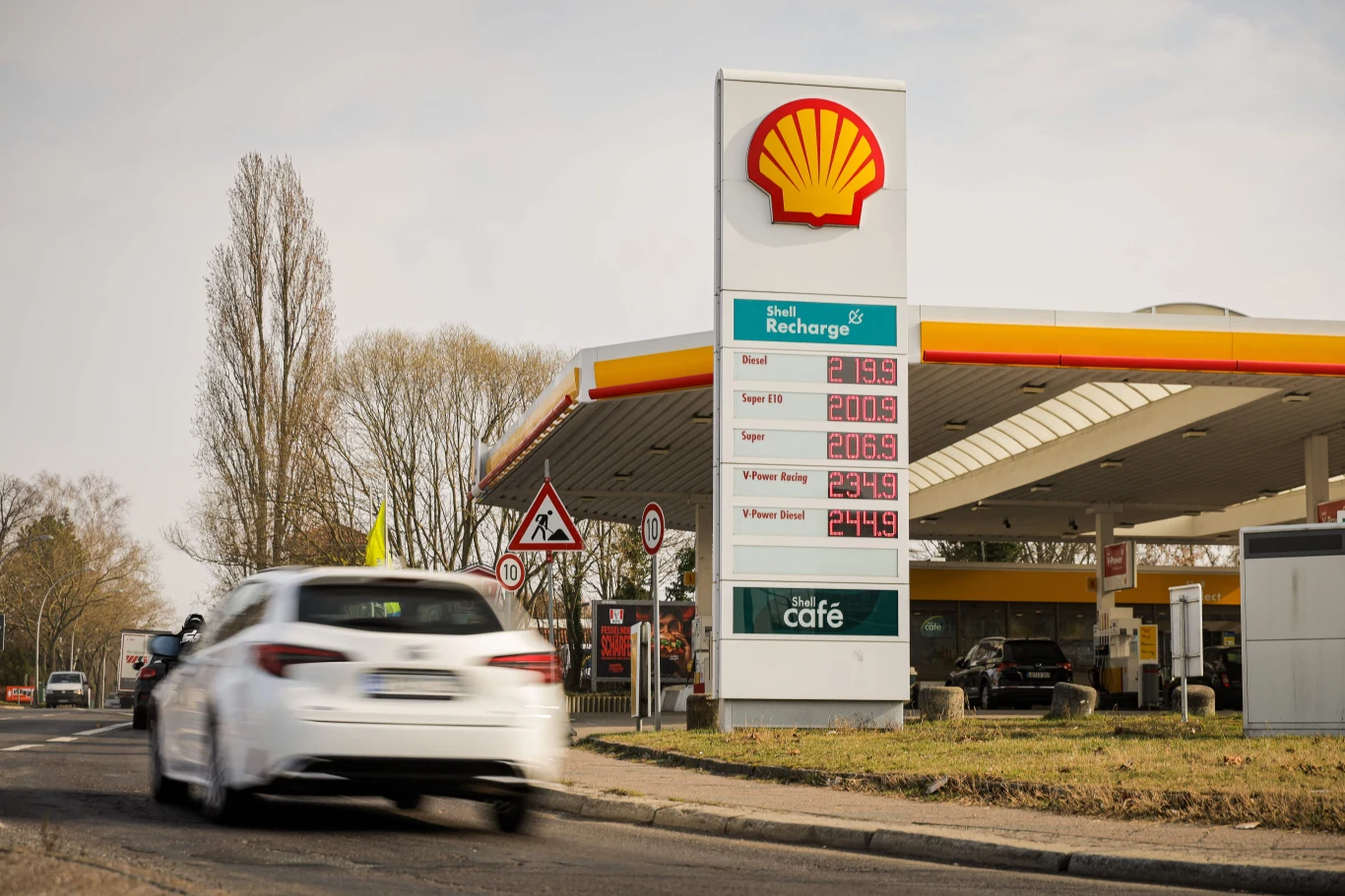 Niemcy zostały szczególnie mocno dotknięte podwyżkami cen paliw Stacja benzynowa Shell, słup z cenami paliw, zaparkowany i nadjeżdżający samochód, drzewa i znaki drogowe w tle.