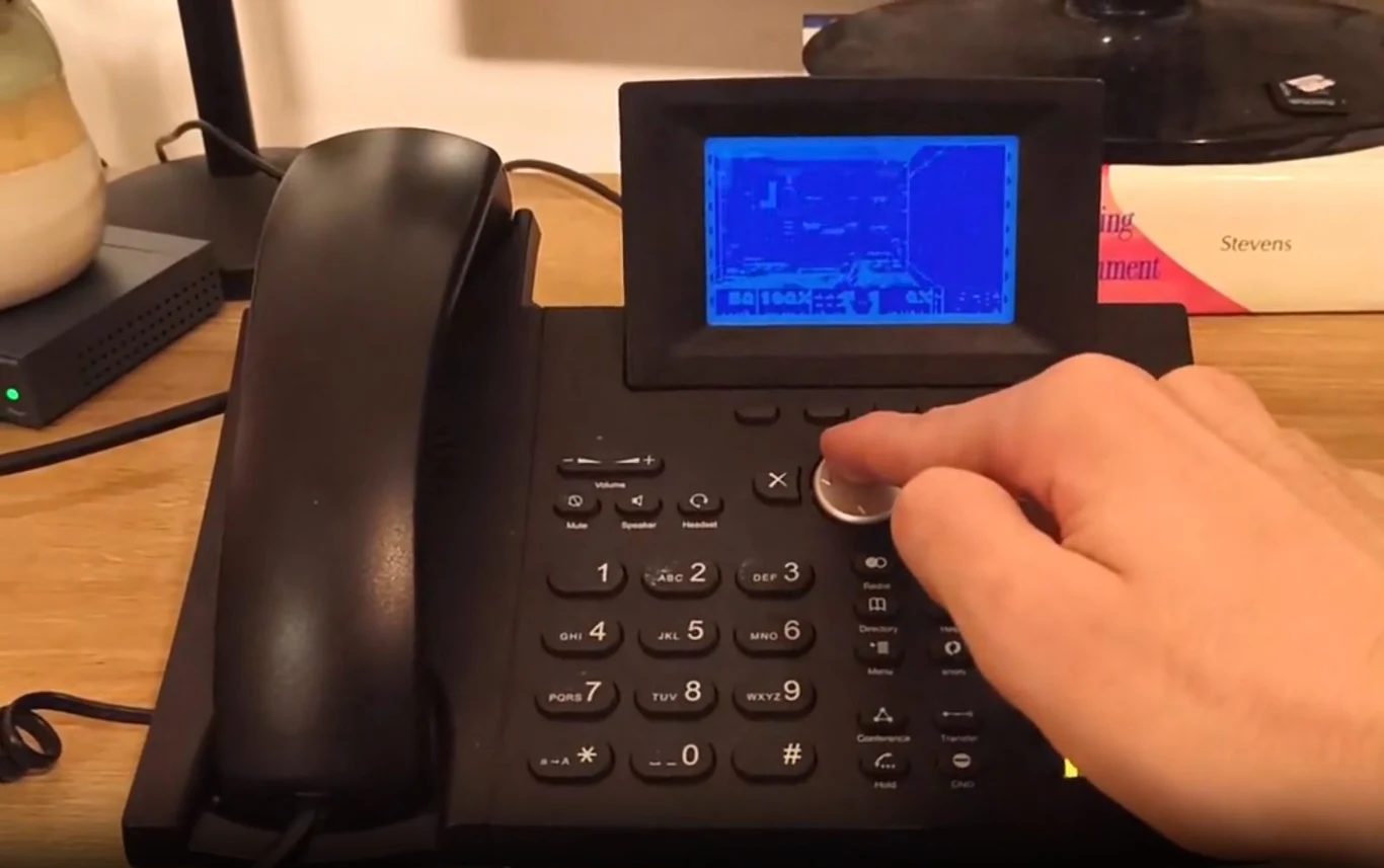 Telefon biurowy z dużym ekranem LCD, przy którym widoczna jest ręka obsługująca pokrętło nawigacyjne; na ekranie wyświetlony jest nieczytelny, niebieski interfejs graficzny.