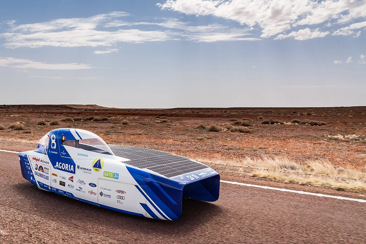 Niebiesko-biały samochód wyścigowy napędzany energią słoneczną jedzie pustą asfaltową drogą przez suchy, pustynny krajobraz pod częściowo zachmurzonym niebem.