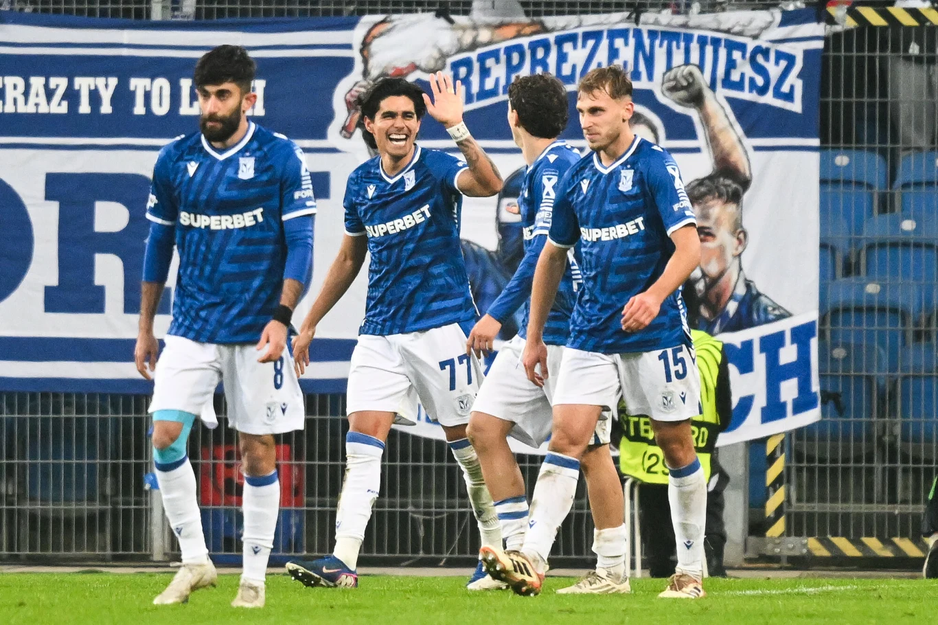 Czterech piłkarzy w niebieskich koszulkach klubu Lech Poznań świętuje zdobytego gola na tle trybun z wywieszonym transparentem i dopingującymi kibicami.