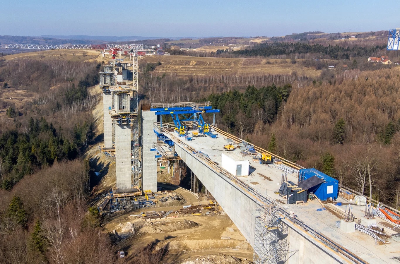 Wysoki most w trakcie budowy nad zalesioną doliną, z widocznymi wieżami podporowymi, maszynami budowlanymi i betonowymi elementami konstrukcyjnymi, otoczony pagórkowatym, leśnym krajobrazem.