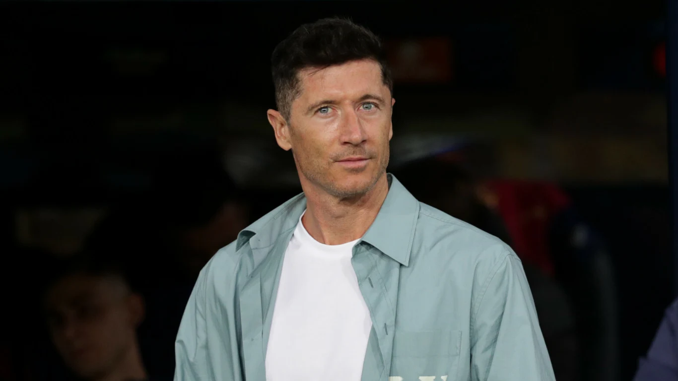 Mężczyzna o ciemnych włosach i niebieskich oczach stoi w półotwartym, jasnozielonym płaszczu na białej koszulce, patrząc spokojnie przed siebie; w tle niewyraźne sylwetki innych osób.