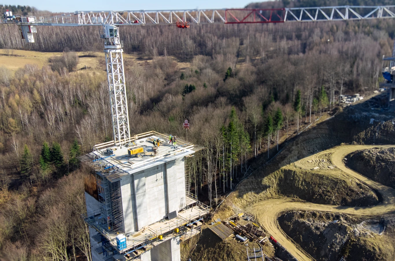 Wysoka betonowa konstrukcja mostowa w trakcie budowy, wyposażona w żuraw wieżowy, otoczona surowym terenem i lasem, w tle widoczna jest metalowa konstrukcja planowanego wiaduktu ponad doliną.